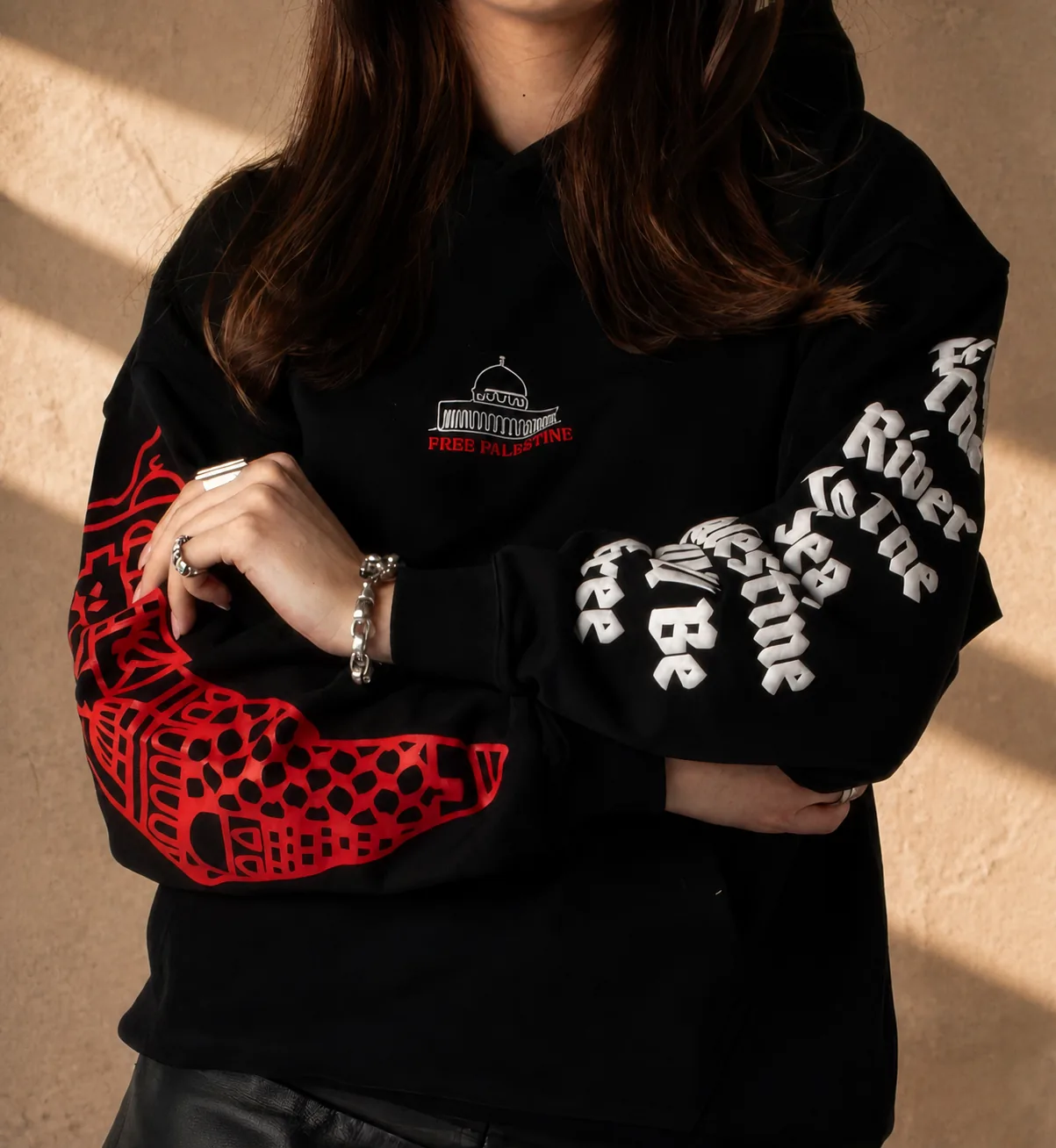 The black Palestine hoodie worn with arms crossed, the Dome and FREE PALESTINE small on the chest, the red map down one sleeve and the gothic text down the other