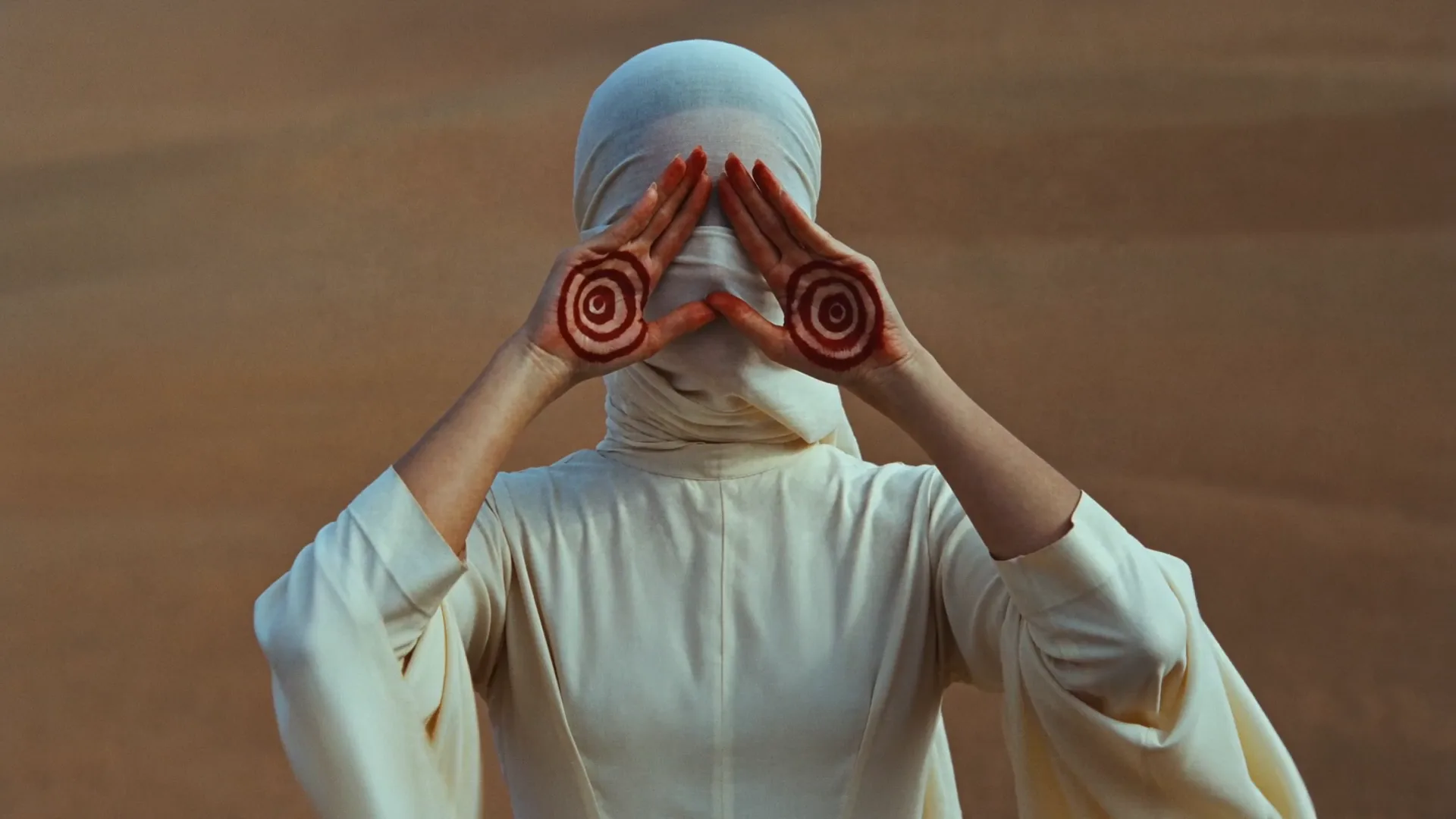 A figure in cream robes in the desert, palms raised over the eyes